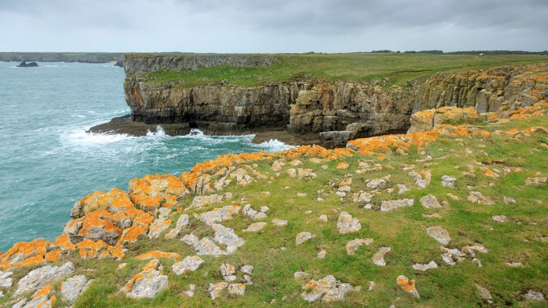 Looking over grass cliff edge to rough sea below with cloudy sky above